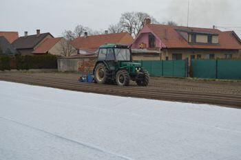 Traktor jedoucí po poli. V pozadí shluk vesnických stavení.